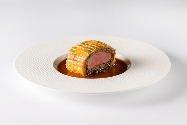 A slice of medium-rare Lamb Wellington wrapped in golden, fluted pastry and served in a rich brown jus on a textured white plate at Kanishka Restaurant.